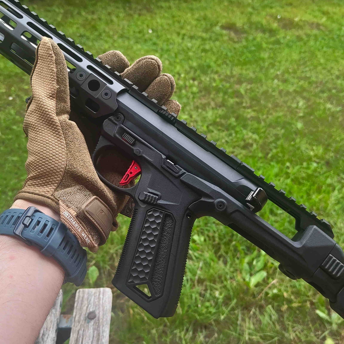 Person holding a black rifle with a red dot sight outdoors on grass